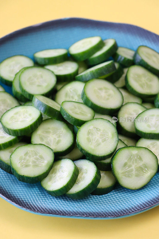 蓝色盘子上的一堆黄瓜(Cucumis sativus)切片的全画幅图像，显示绿色的皮肤和白色的果肉，白色的种子，黄色的背景，高架视图，重点放在前景上